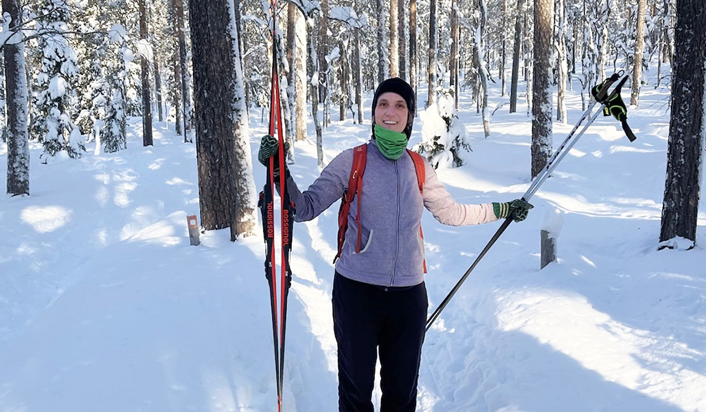 Tiina Vainio-Kaila står i en snöig skog och håller ett par skidor i ena handen och stavarna i den andra.