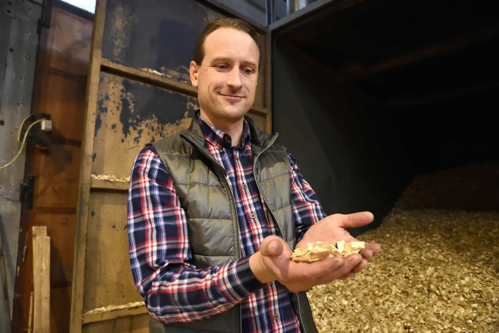 Carbofex verkställande direktör Sampo Tukiainen håller i skogsflis som används i tillverkningen av biokol. Bakom honom syns en stor hög med flis.