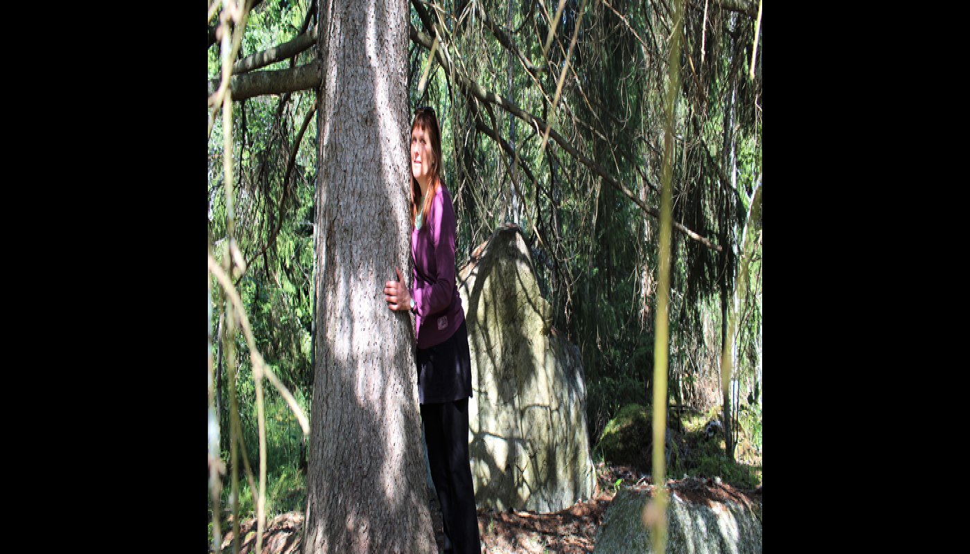 Kirsi Salonen står och lutar sig mot en trädstam, en gammal gran. Solstrålar sipprar in mellan grenverken.