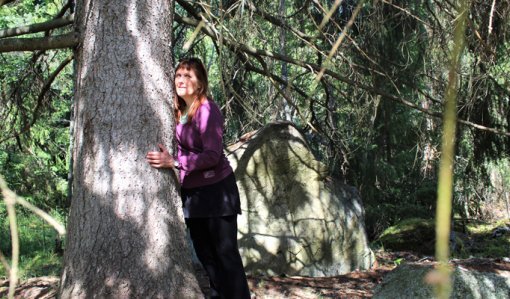 Kirsi Salonen står och lutar sig mot en trädstam, en gammal gran. Solstrålar sipprar in mellan grenverken.