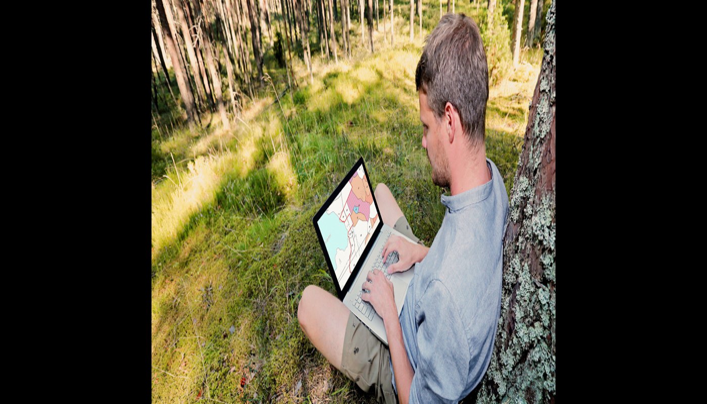  En man sitter på en gräsmatta med en dator i famnen och tittar på information om sin egen skog i e-tjänsten MinSkog.fi. Han lutar sig ryggen mot ett träd. Runt honom syns gräsmatta och i bakgrunden skog.