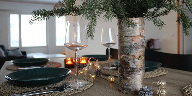 Juldukning. På bordet står tallrikar, bestick, vinglas, två värmeljus, prydnadsbollar och prydnadskottar. I förgrunden står en björkvas med barrträdskvistar.