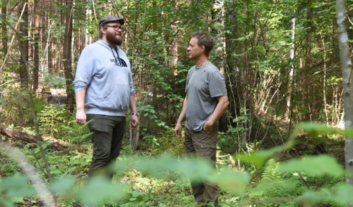 Skogsägaren Tero Ahava och naturvårdsexperten Jukka Ruutiainen står i hassellunden och diskuterar. Växtligheten runtomkring dem är grön och frodig.