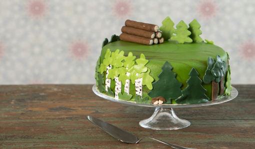 Tårtan är helt täckt av grön marsipan och garnerad med gran- tall- och björkfigurer av marsipan längs sidorna. Uppe på kakan ligger en timmerhög av marsipan, och bakom den står några marsipangranar.