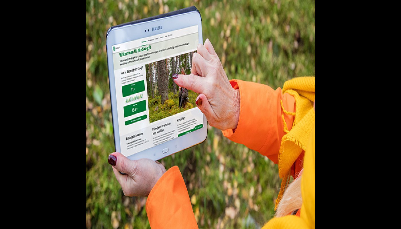 En person håller i en surfplatta där förstasidan av tjänsten MinSkog.fi är öppen. Hen pekar med fingret på förstasidan. 