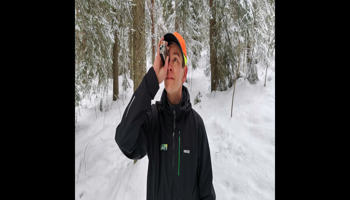 Skogsrådgivaren Ari Kasanen står i en snöig skog och håller en höjdmätare framför ögat.