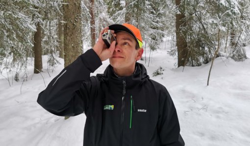 Skogsrådgivaren Ari Kasanen står i en snöig skog och håller en höjdmätare framför ögat.