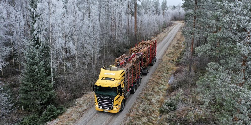 En virkestransportbil har fotograferats uppifrån. Fordonet har gul för och är fullastat. Skogsvägen som fordonet kör på kantas av skog.