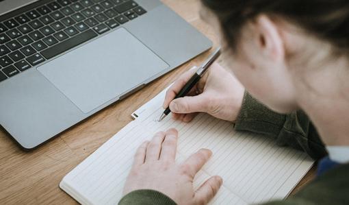 En person skriver anteckningar i ett häfte. Bredvid på bordet finns en bärbar dator.
