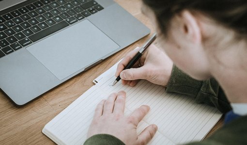 En person skriver anteckningar i ett häfte. Bredvid på bordet finns en bärbar dator.
