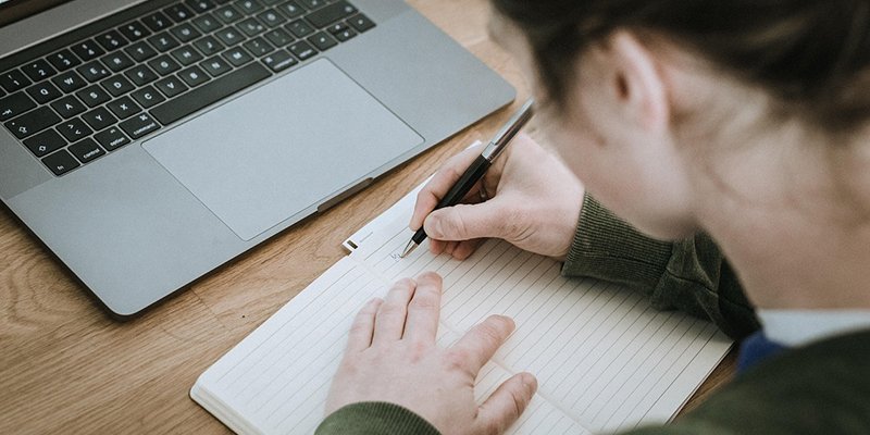 En person skriver anteckningar i ett häfte. Bredvid på bordet finns en bärbar dator.
