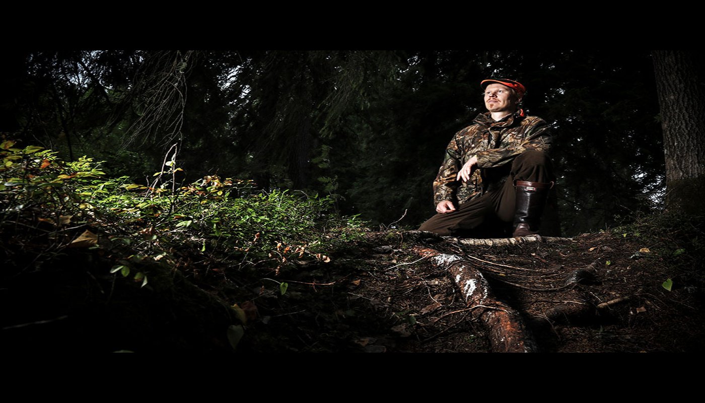 Viltplaneraren Teemu Lamberg sitter med ena knät i marken vid rötterna av ett träd. Skogen bakom honom är mörk.