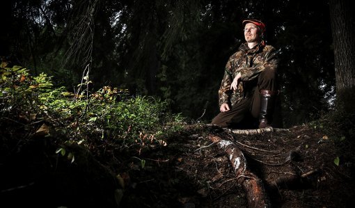 Viltplaneraren Teemu Lamberg sitter med ena knät i marken vid rötterna av ett träd. Skogen bakom honom är mörk.