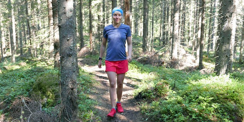 Saara Grönholm kommer leende gående emot kameran på en stig. Runt henne syns granar och risvegetation, och solen lyser in mellan träden.