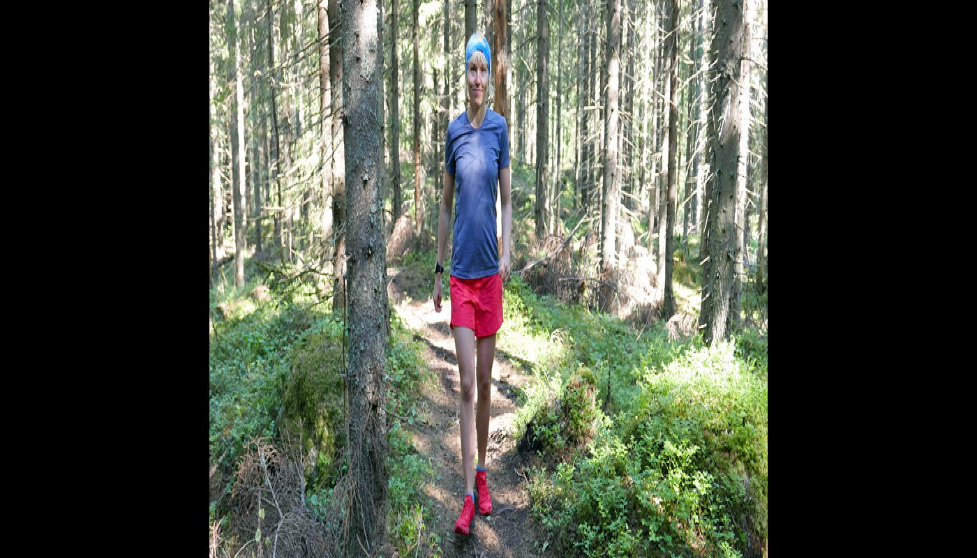 Saara Grönholm kommer leende gående emot kameran på en stig. Runt henne syns granar och risvegetation, och solen lyser in mellan träden.