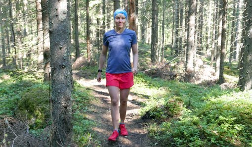 Saara Grönholm kommer leende gående emot kameran på en stig. Runt henne syns granar och risvegetation, och solen lyser in mellan träden.