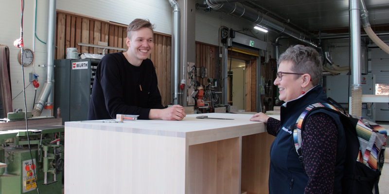 Miikka Kotilainen står bakom ett högt bord av trä och pratar med företagsrådgivare Leila Laukkanen i Puuartistis produktionslokal.