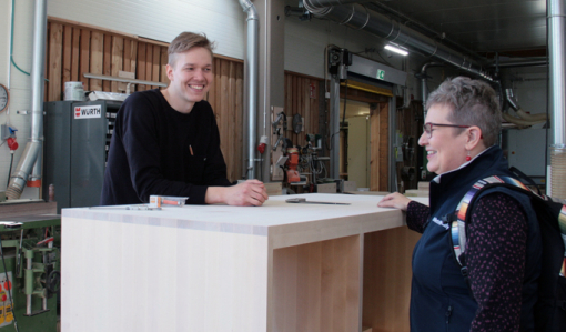 Miikka Kotilainen står bakom ett högt bord av trä och pratar med företagsrådgivare Leila Laukkanen i Puuartistis produktionslokal.