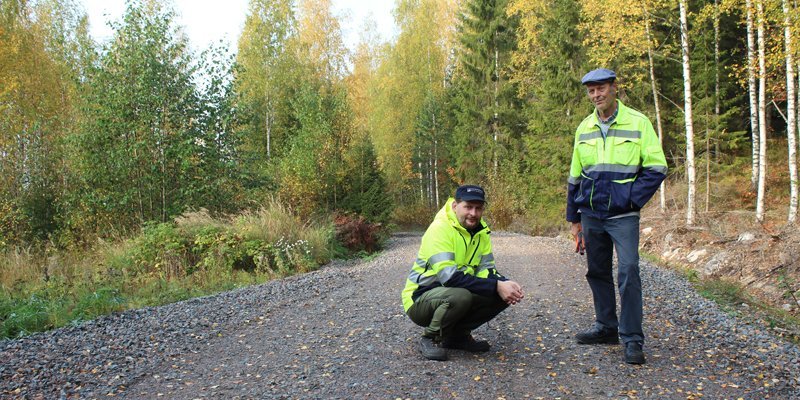 Tomi Vähä-Kouvola sitter på huk på en skogsväg. Bredvid honom står Tapio Kahilaniemi. Gula löv har fallit på vägen, som omges av skog.
