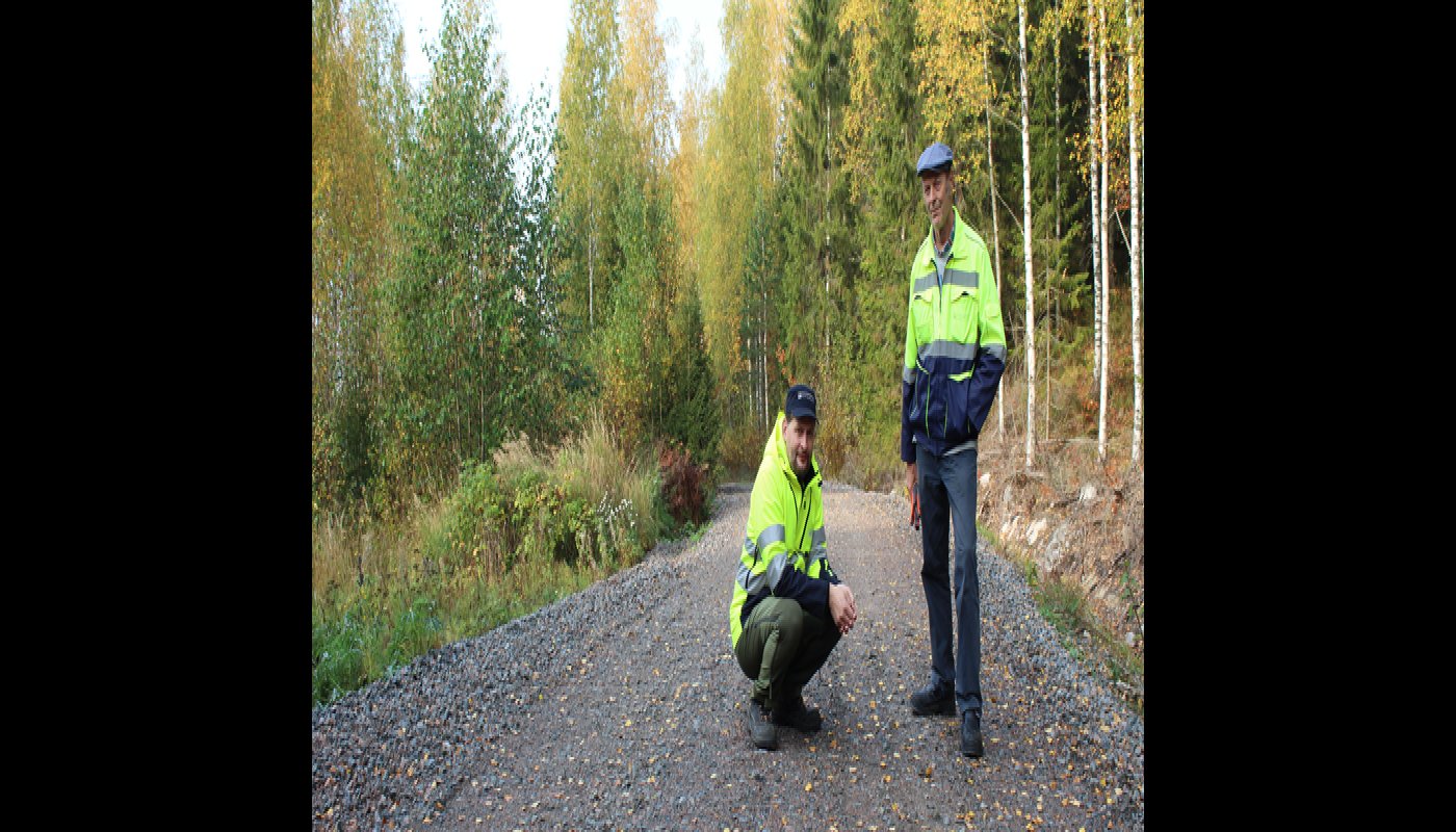 Tomi Vähä-Kouvola sitter på huk på en skogsväg. Bredvid honom står Tapio Kahilaniemi. Gula löv har fallit på vägen, som omges av skog.