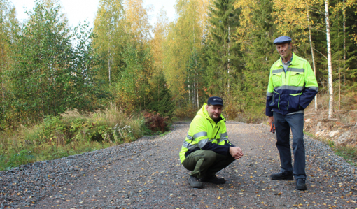 Tomi Vähä-Kouvola sitter på huk på en skogsväg. Bredvid honom står Tapio Kahilaniemi. Gula löv har fallit på vägen, som omges av skog.