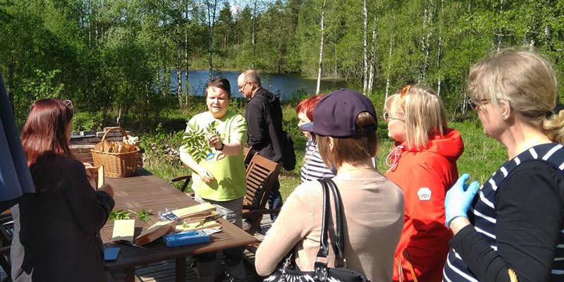 Producenten Leena Löf står och håller upp vilda örter inför en grupp åhörare. Bakom henne syns björkar och en sjöglimt.