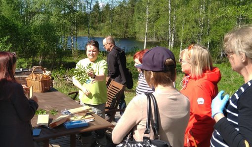 Producenten Leena Löf står och håller upp vilda örter inför en grupp åhörare. Bakom henne syns björkar och en sjöglimt.