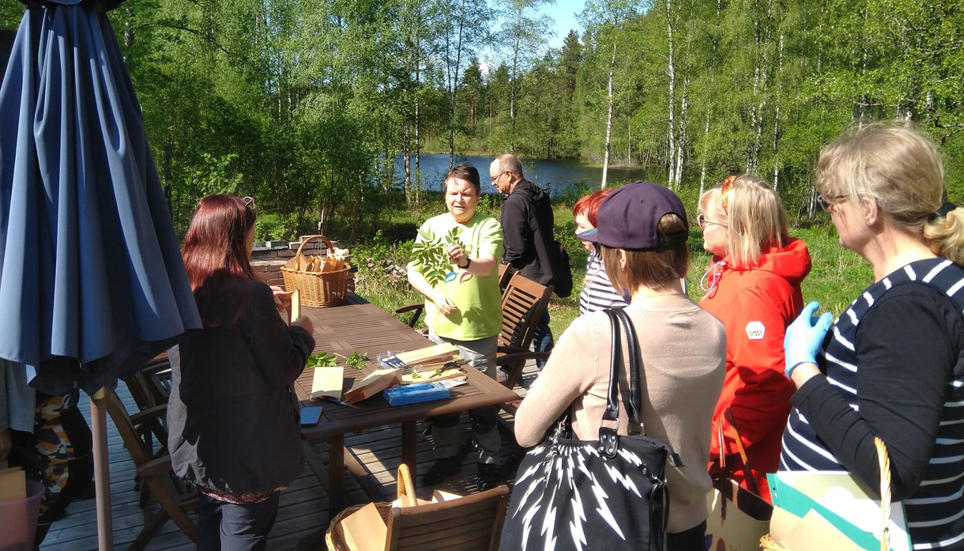 Naturproducenten Leena Löf presenterar vilda örter för åhörarna runt omkring henne. I bakgrunden finns björkar och en sjöutsikt.