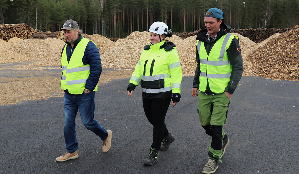 Företagaren Jarmo Hieto, energibolaget Loimuas operativa chef Karoliina Kärkäs och jord- och skogsbruksföretagaren Lauri Tapola vid en virkesterminal. Bakom dem syns högar av flis och oflisat virke.