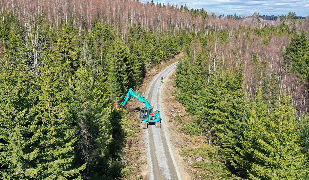 En grävmaskin på en skogsväg, fotograferad från luften. Påp den har man monterat avverkningsutrustning och tar bort träd vid vägkanten. en person står och ser på.