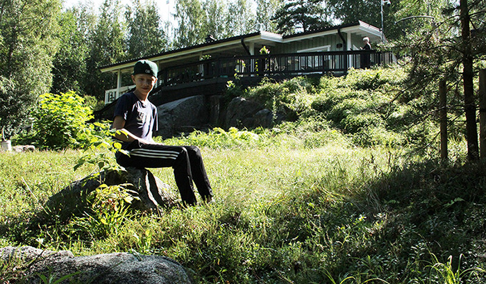 Sampo Riikonen istuu kivellä Kantosen mökkipihalla, johon on asennettu kunttaa. Taustalla on mökki. 