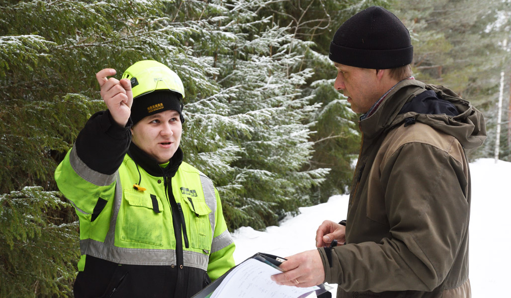 Skogsmaskinsföraren Mikko Inkinen och Jukka Ruutiainen står och samtalar på en skogsväg. Bakom dem syns snöiga granar.