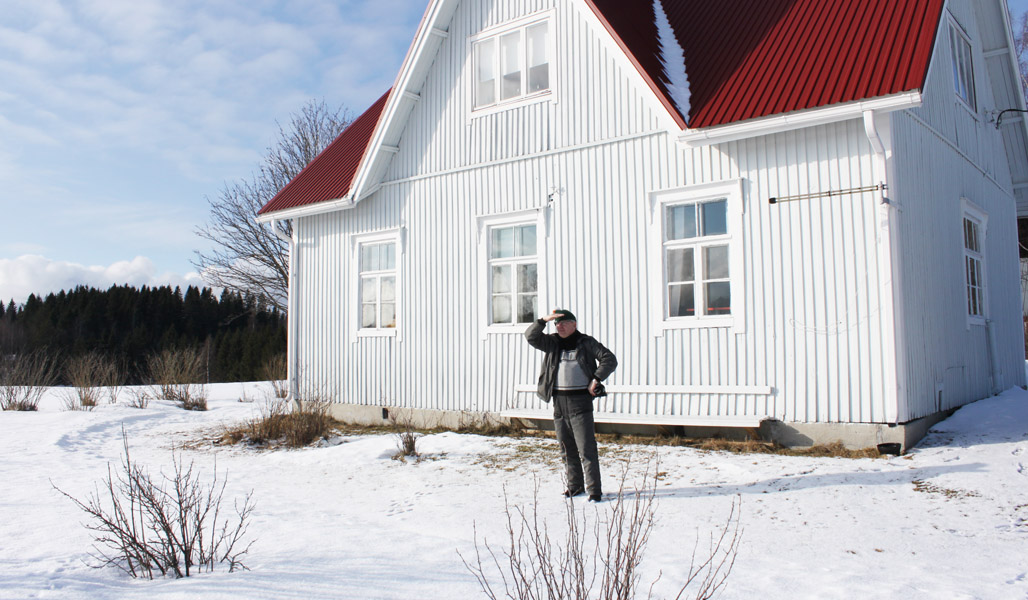 Timo Nissinen står framför huset som är släktens sommarviste och spanar ut mot skyddsobjektet. Det är snö på marken, i bakgrunden syns buskar på gården och längre bak skog.