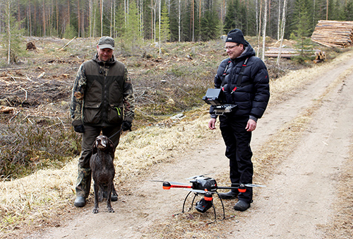 Tommi Kinnunen, hunden Heta och Video Drone Oy:s Juhani Mikkola står på en skogsväg intill en avverkningsyta. Mikkola har en värmekamera hängande runt halsen och på vägen står en drönare.