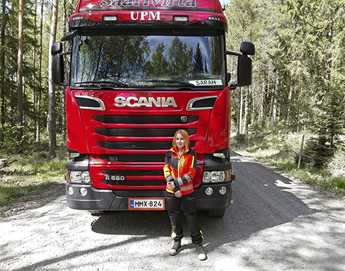 Sarah Lindahl som kör virkestransporter står framför en röd virkestransportbil på en skogsväg.