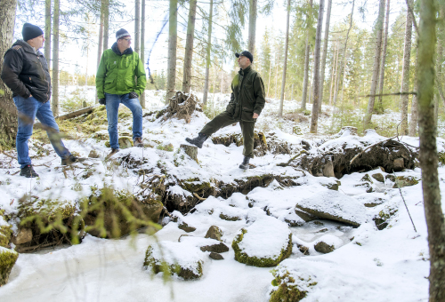 Visa Mikkola, Pentti Pakarinen och Mika Tuomola står och pratar invid en bäck i naturtillstånd. Det är snö på marken.