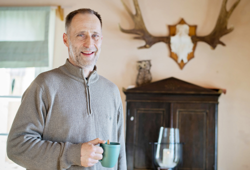 Skogsägaren Mika Tuomola stående i sitt hem med kaffekopp i handen. Vid väggen bakom honom står ett brunt skåp och på det en prydnadsuggla. På väggen hänger ett par älghorn.