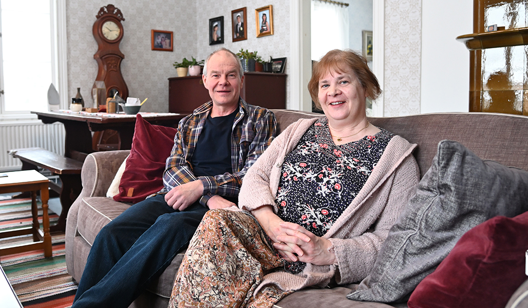 Tapani och Virpi Isotalo sitter i vardagsrumssoffan. Bakom dem vid väggen står ett golvur, en kakelugn och ett timrat bord. På väggen hänger studentfoton på parets barn.