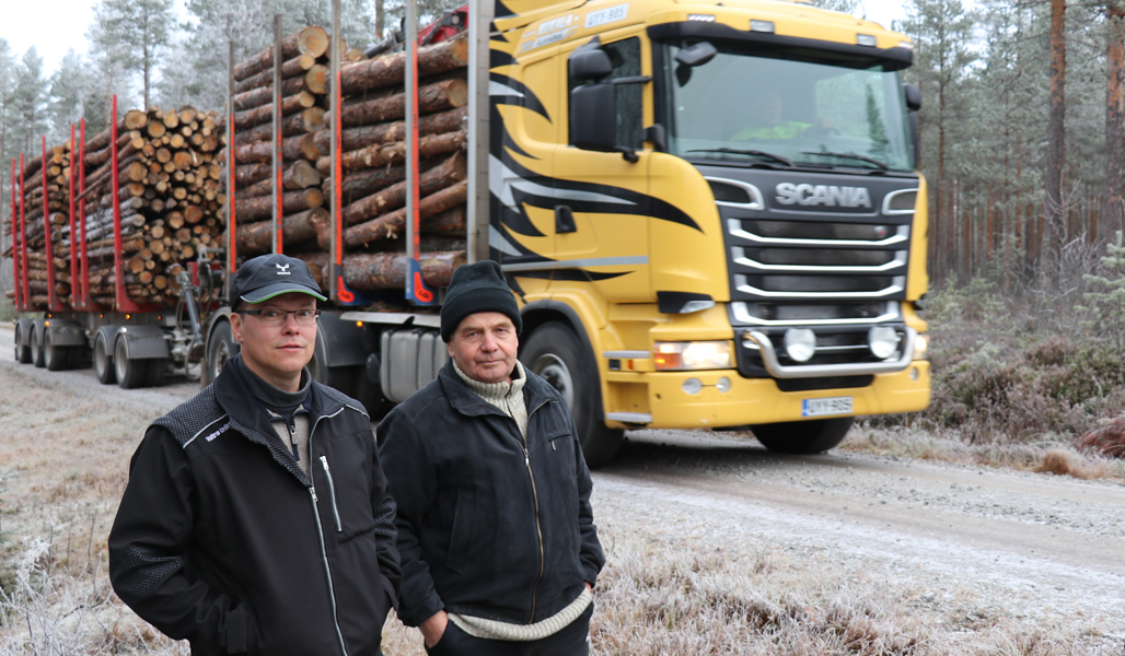 Jouni Mäkiluoma till vänster, ordförande för väglaget, och Juhani Kosola, medlem i bestyrelsen står på en väg med en virkestransportbil bakom sig.