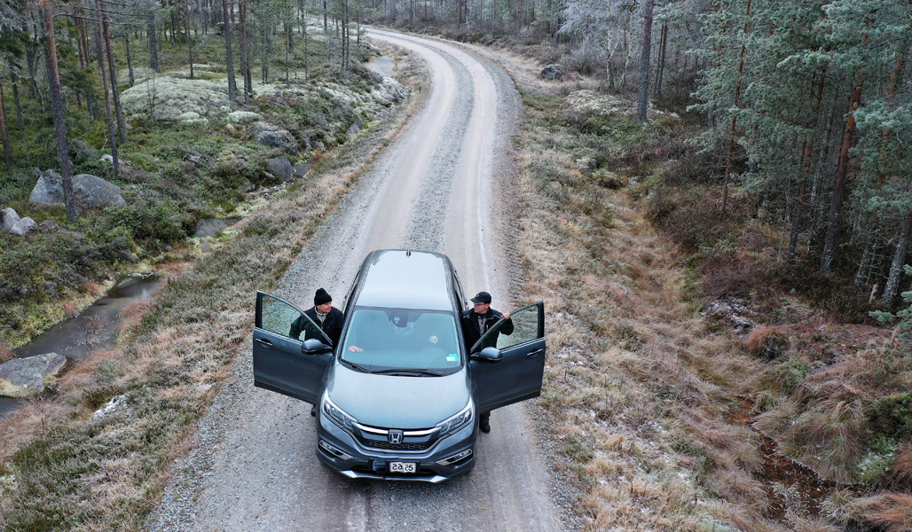 En bil har fotograferats uppifrån på en skogsväg. Två män stiger ut ur bilen.