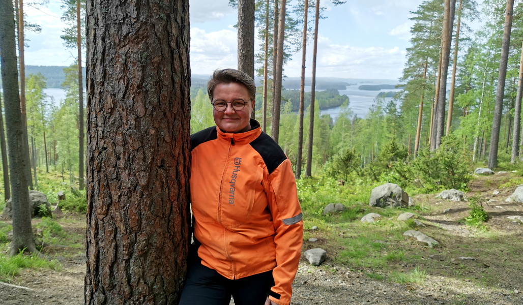 Anne Ruokonen står bredvid en tall. Bakom henne syns skog och ett insjölandskap.