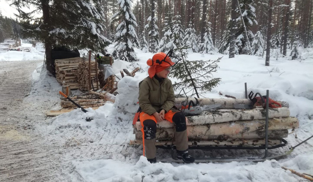 Pirjo Paldanius sitter klädd i skogsarbetarens skyddskläder på en timmertrave i skogen på vintern. 