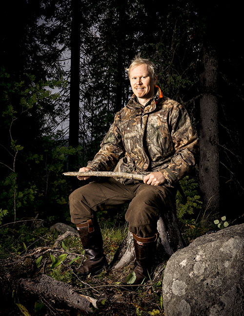 Viltplaneraren Teemu Lamberg sitter på en stubbe och ler. I händerna håller han en liten trädgren. Skogen bakom honom är mörk.