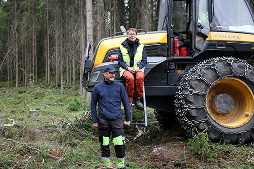 Skogsmaskinsförare Kyösti Karhila och Teija Hyytiäinen-Koskimäki framför en skogsmaskin i skogen.