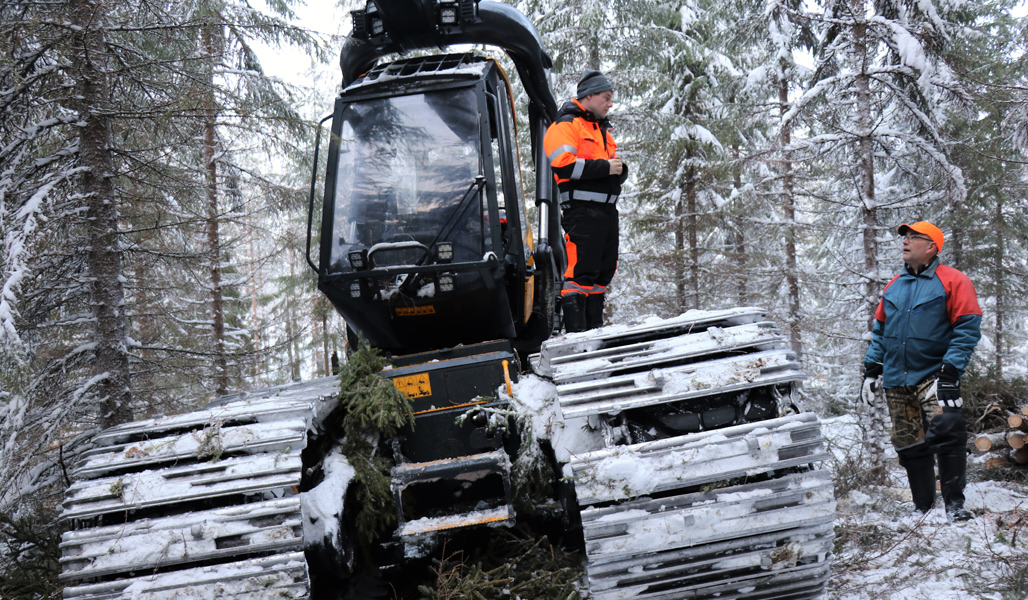 Skogsmaskinföraren Mikko Ahonen står på maskinen och diskuterar med Mikko Tiirola i en snöig skog.