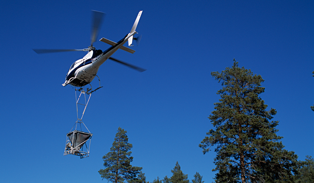 En flygande helikopter mot en blå himmel. Under helikoptern hänger kärlet med askgödsel som sprids i skogen.